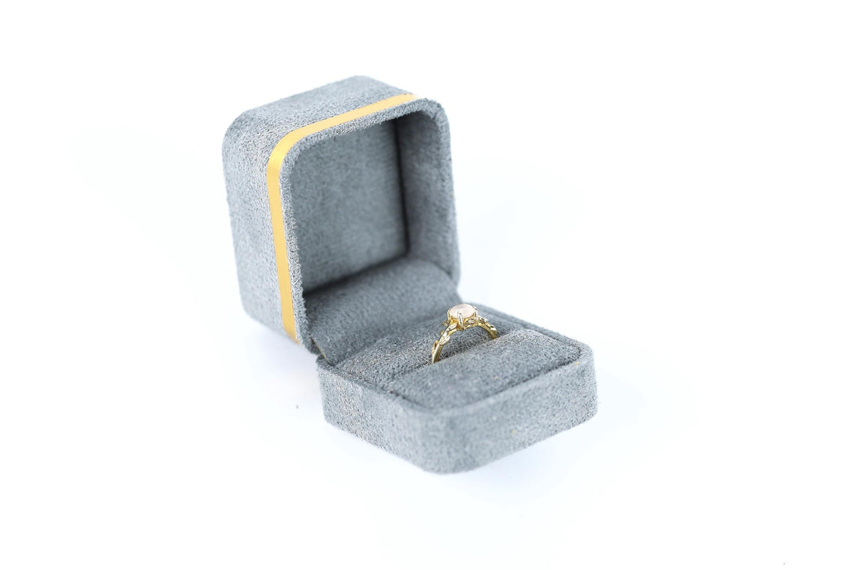 A gold ring with a diamond set in a gray velvet jewelry box with a yellow stripe.