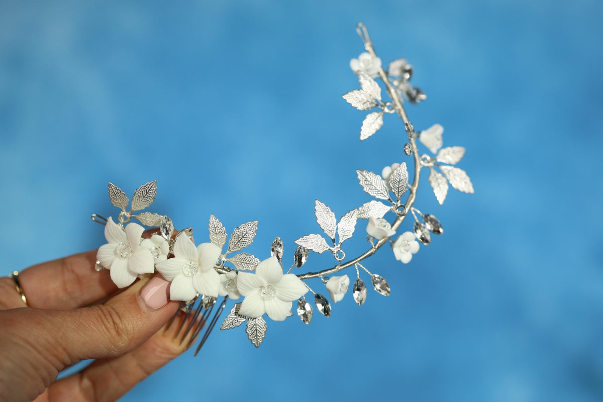 A hand holding a decorative hair clip with white flowers and leaves against a blue background.