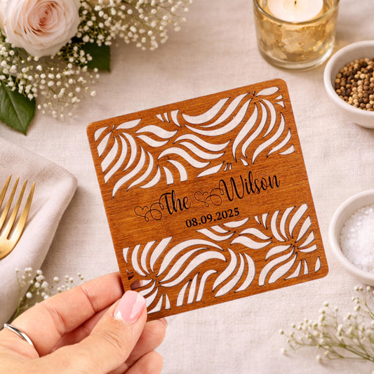 A hand holding a wooden card with a floral design and text on it, surrounded by other decorative items such as flowers, a candle, and a bowl of spices.
