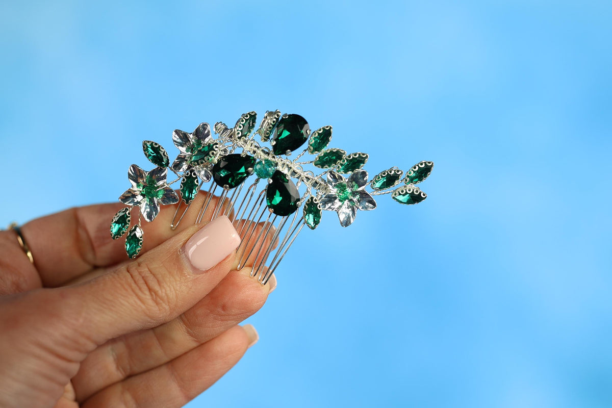 A hand holding a decorative hair accessory with green and silver gemstones against a blue background.