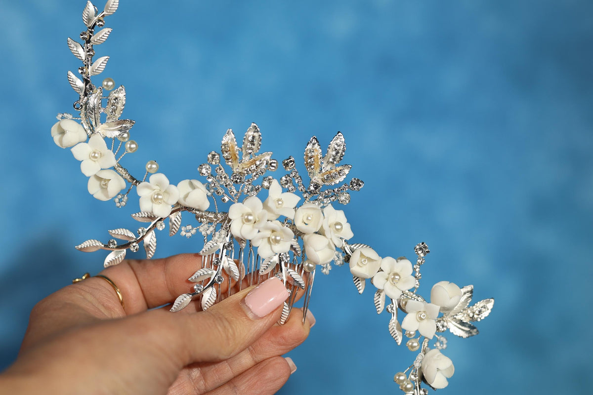 A hand holding a decorative hair accessory with white flowers and leaves against a blue background.