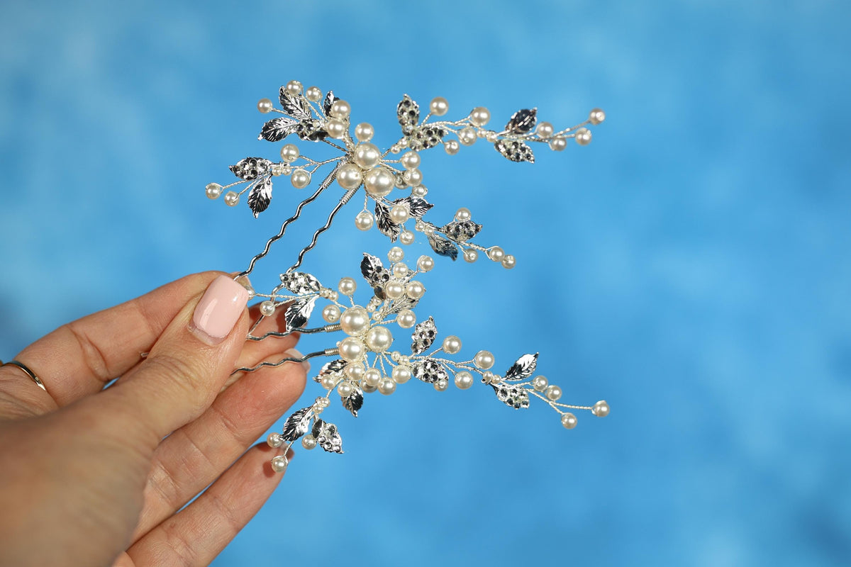 A hand holding a decorative hair clip with small pearls and leaves against a blue background.