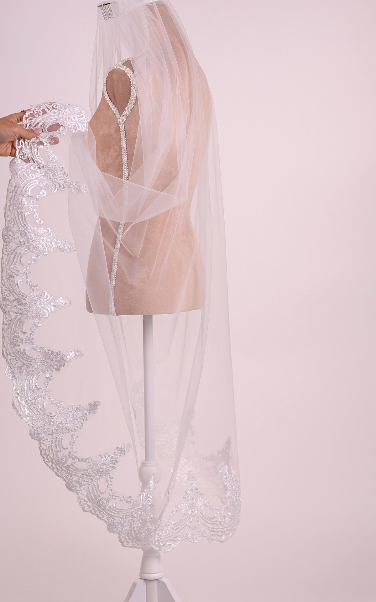 A white wedding veil with a delicate lace-like pattern is displayed on a mannequin head, set against a light pink background.
