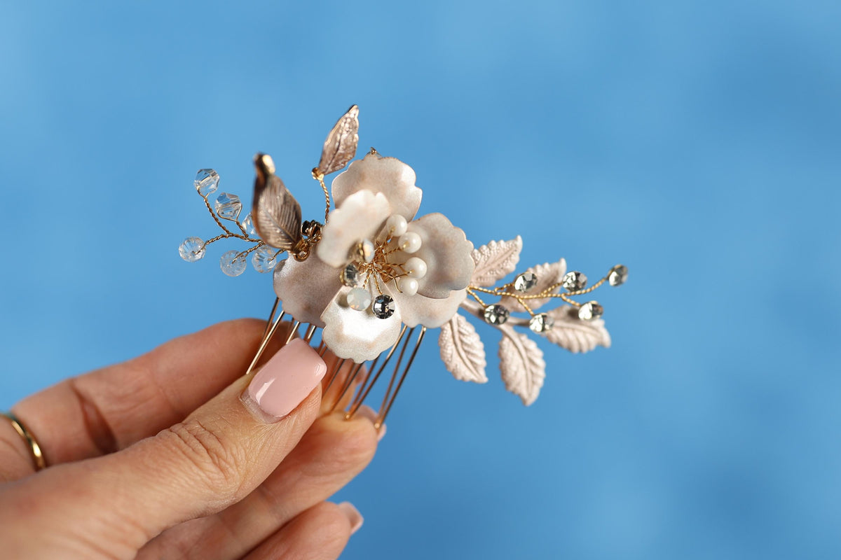 A hand holding a decorative hair clip with a flower and leaves against a blue background.
