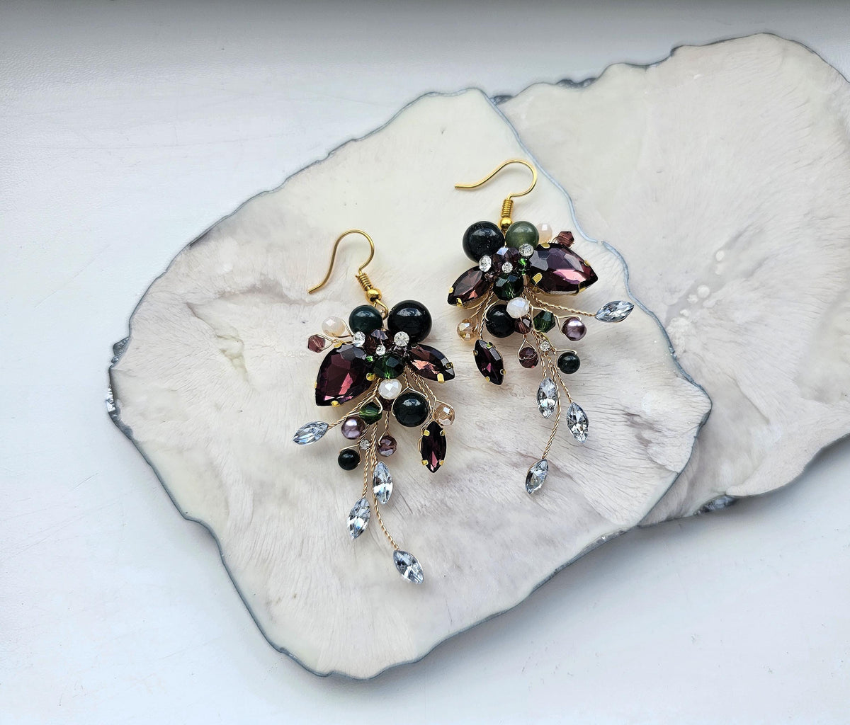A pair of elegant earrings with a floral design and various gemstones, including purple, green, and clear stones, are displayed on a white surface.