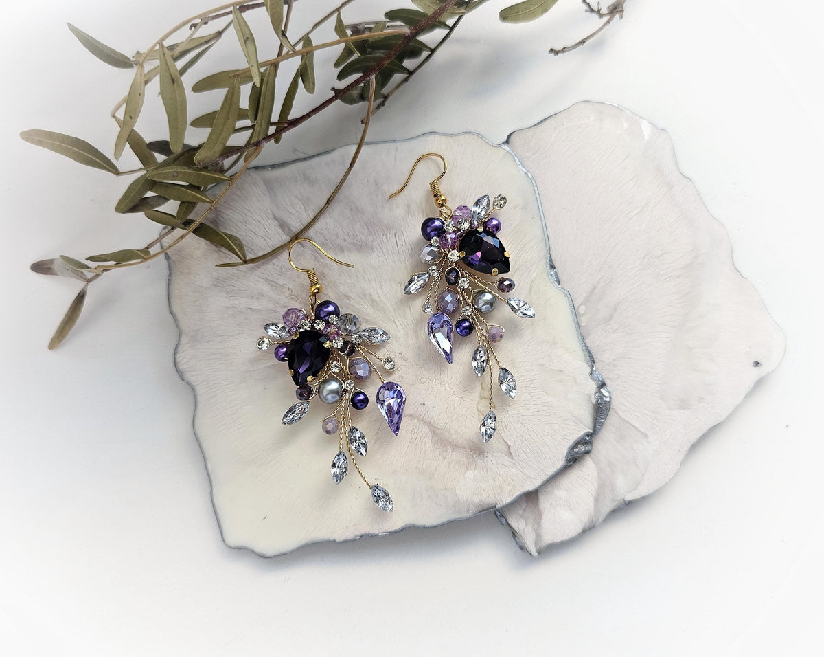 A pair of elegant earrings with purple and blue gemstones, placed on a white surface alongside some green leaves.