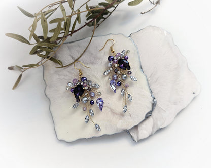 A pair of elegant earrings with purple and blue gemstones, placed on a white surface alongside some green leaves.