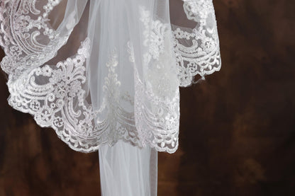 A close-up view of a delicate white lace veil with intricate floral patterns, hanging against a dark background.