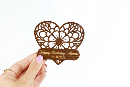 A hand holding a wooden heart-shaped object with a personalized message on it.