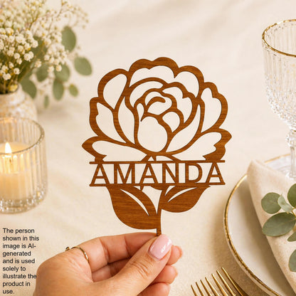 A hand holding a wooden rose-shaped cake topper with the name "Amanda" written on it, placed on a table with a candle, glassware, and greenery.