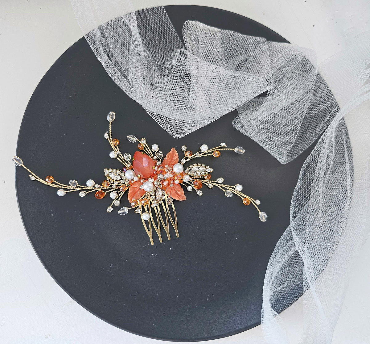 A decorative hair clip with a floral design and sparkling gemstones rests on a black plate, accompanied by a white net or veil.
