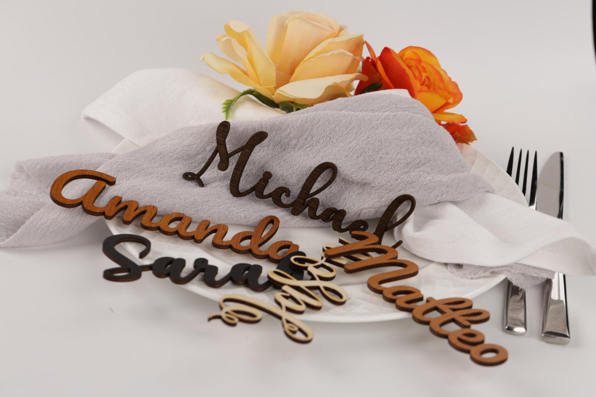 a table setting with a white plate, a napkin, and a floral arrangement. The table setting includes wooden name cards with names on them, as well as a fork and knife.