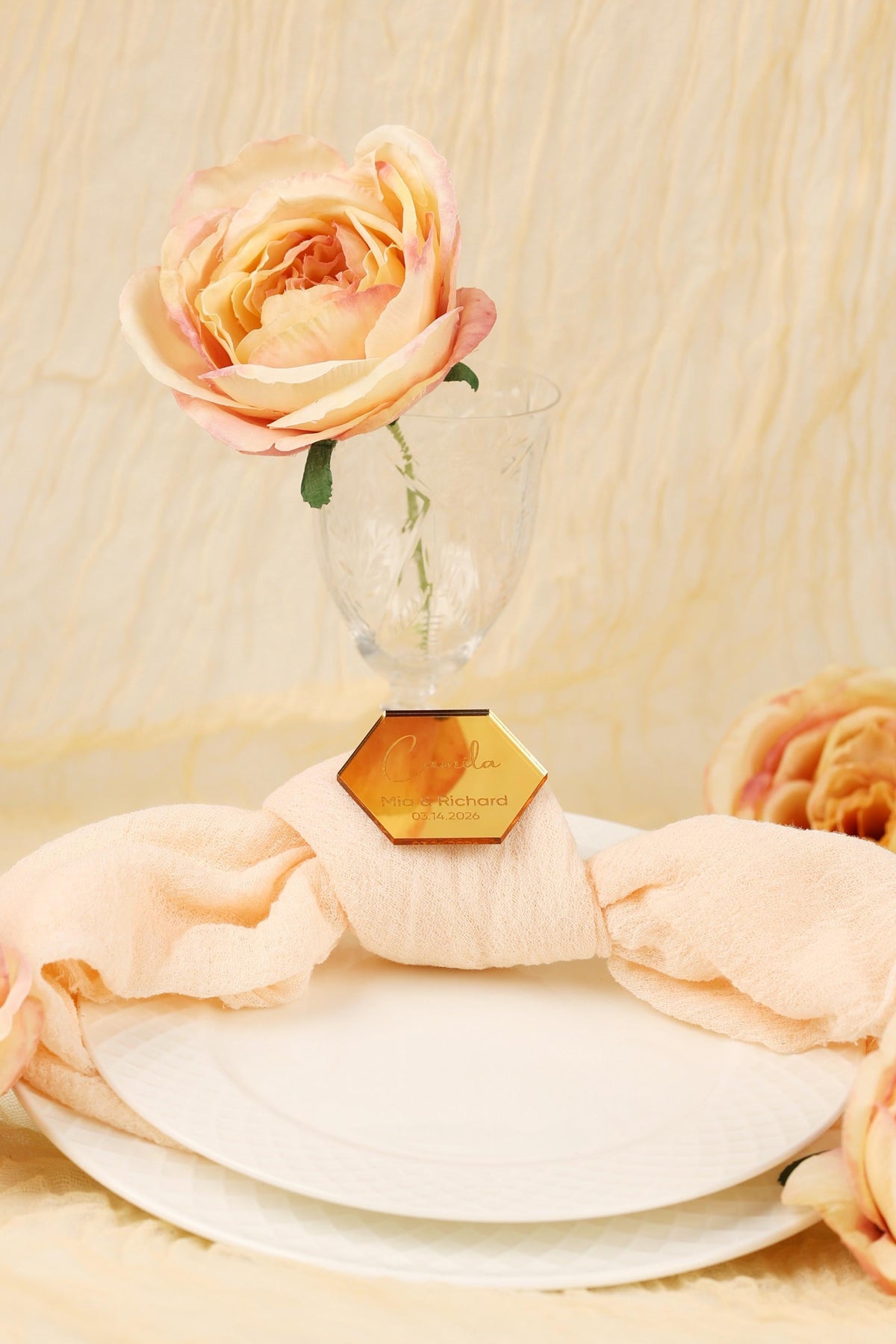 A single pink rose in a glass vase is placed on a white plate, with a gold hexagonal tag attached to the plate.