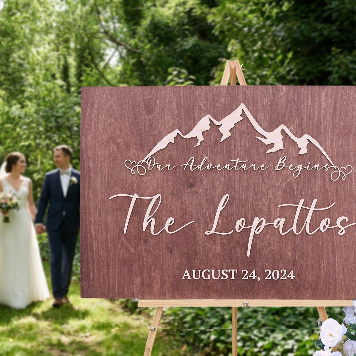 A wooden sign with the text "Our Adventure Begins" and "The Lopatos" is displayed in a grassy area, with a couple walking in the background.