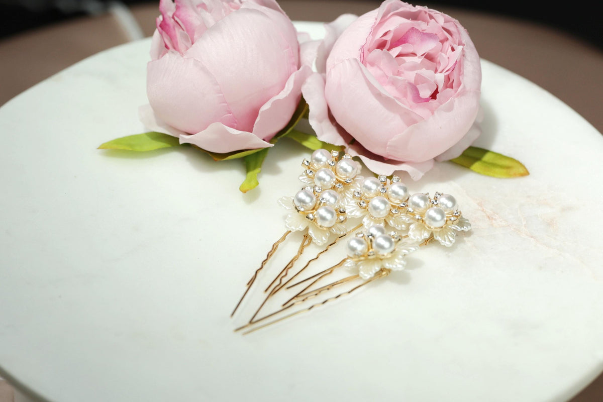 A white marble surface displays a pair of pink peonies and a set of gold and white pearl hair clips.
