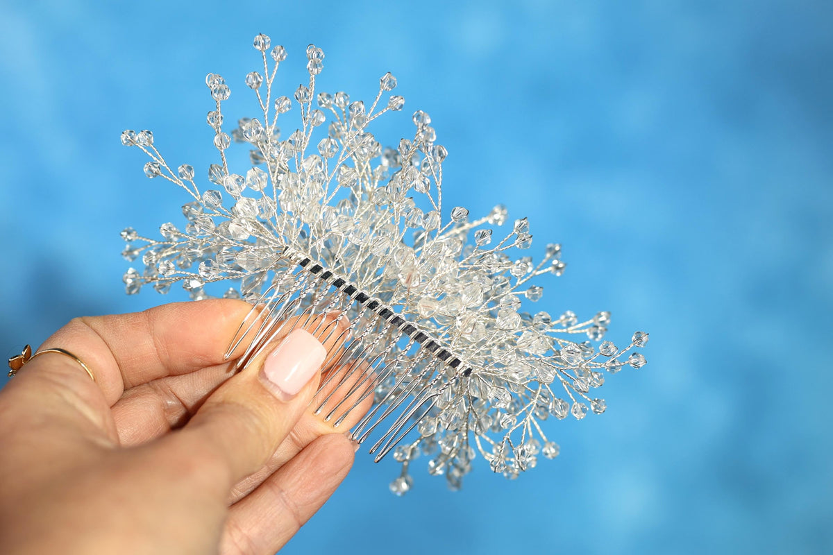 A hand holding a silver hairbrush adorned with small, clear crystal branches against a blue background.