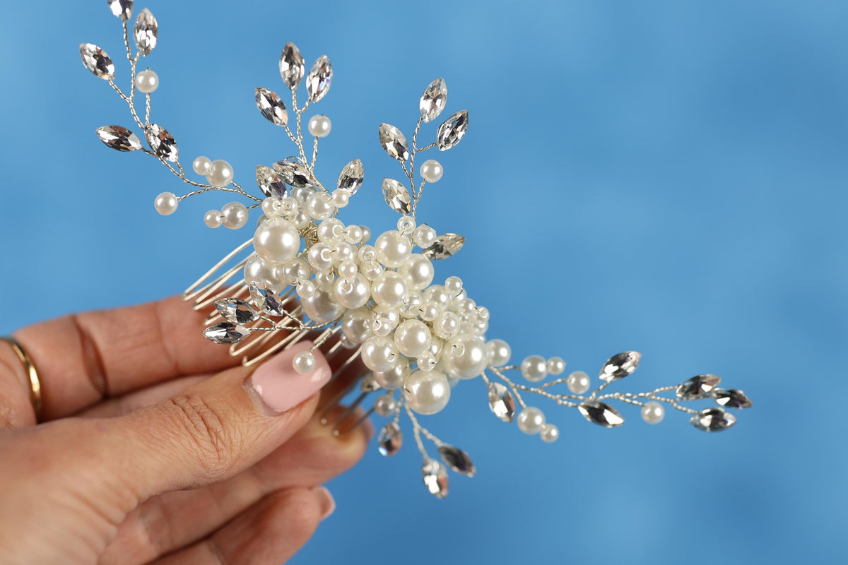 A hand holding a decorative hair accessory made of white pearls and silver beads against a blue background.