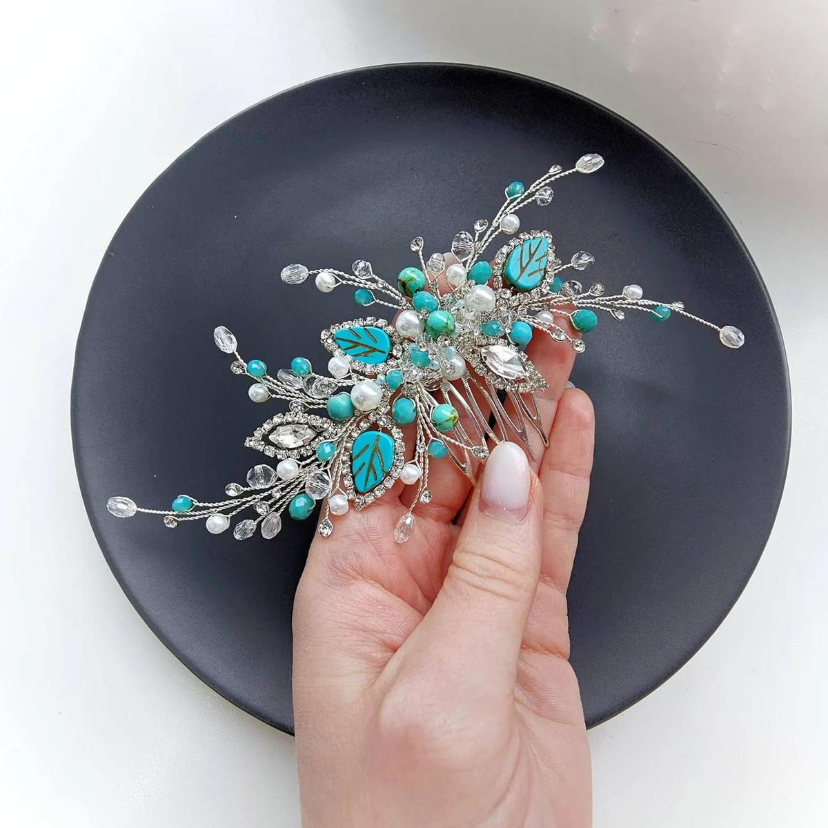 A hand holding a decorative hair accessory with turquoise and white elements, such as leaves and beads, against a black background.