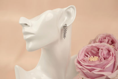 A white mannequin head wearing a pair of dangling earrings, with a bouquet of pink roses in the background.