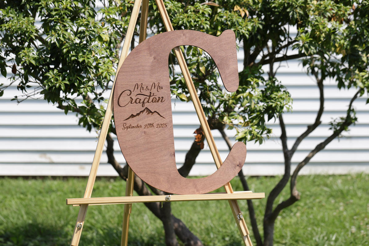 A wooden sign with the letter 'C' on it, displaying the text "Mr. & Mrs. Craftan September 20th 2015" in a garden setting with trees in the background.