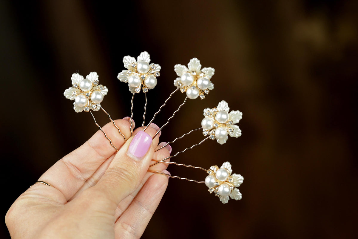 A hand holding a group of six white pearl hair clips with gold accents, arranged in a fan-like pattern.