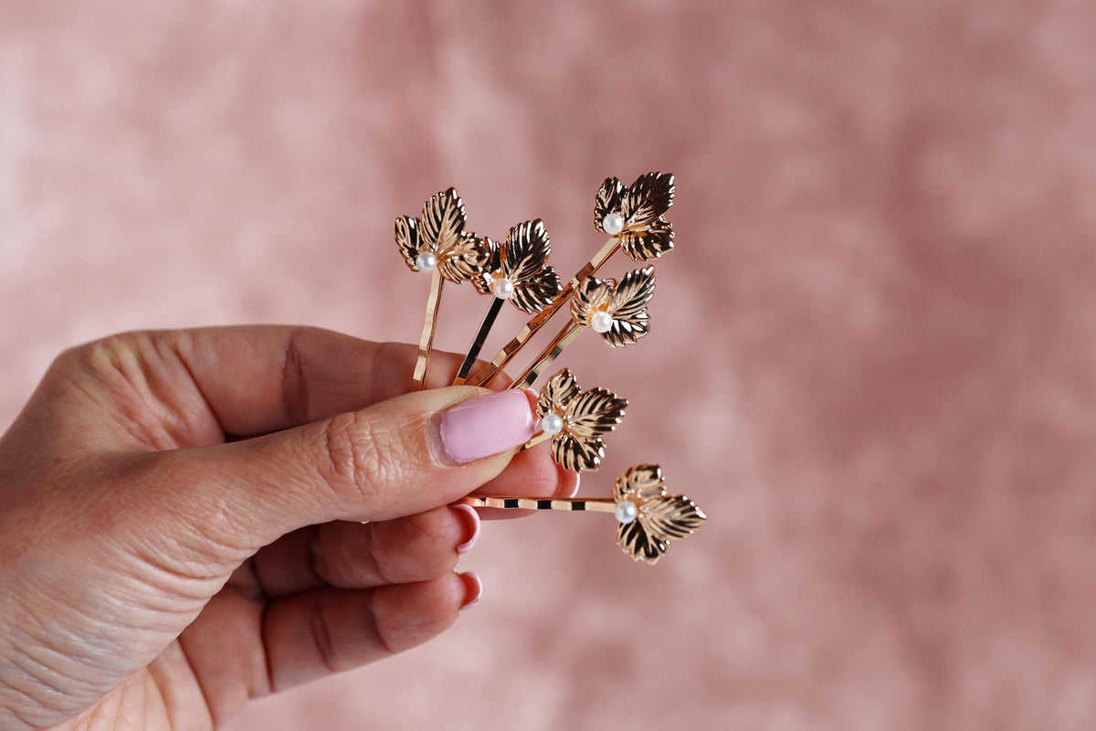 A hand holding a small group of gold leaf-shaped hair clips against a pink background.