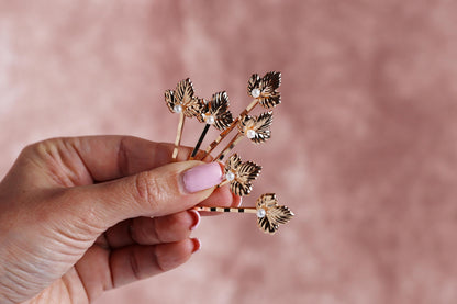 A hand holding a small group of gold leaf-shaped hair clips against a pink background.