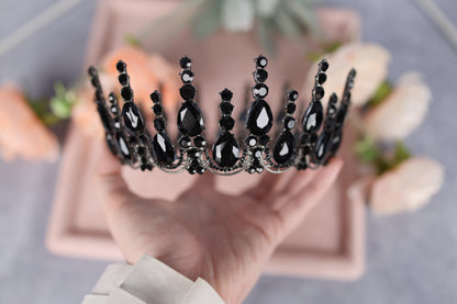 A hand holding a black tiara adorned with black gemstones against a blurred background of pink flowers.