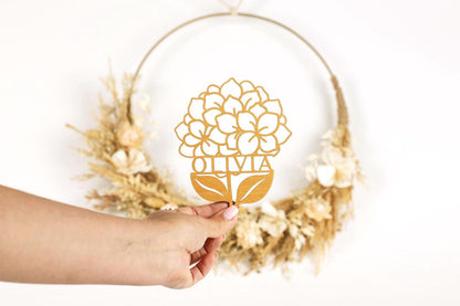 A hand holding a paper cutout of a flower with the name 'Olivia' written on it, in front of a wreath made of dried flowers.