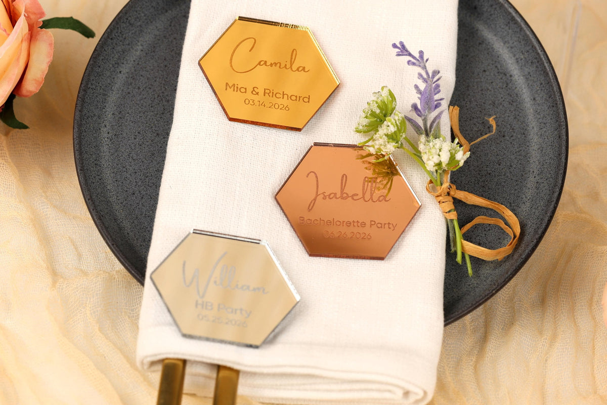 a table setting with a white napkin, a black plate, and three hexagonal glass or metal tags with personalized names and dates.