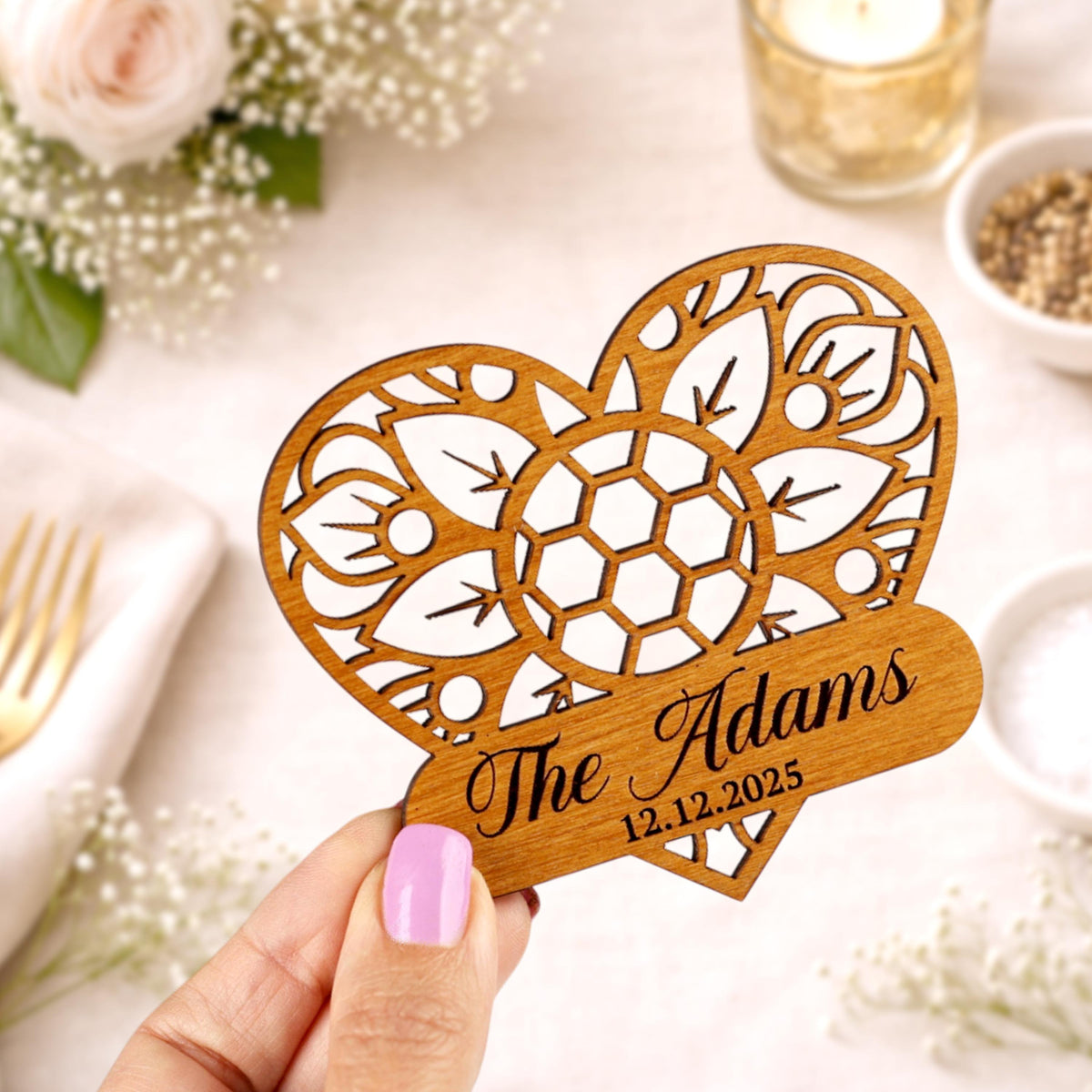A wooden heart-shaped object with a floral design and the text "The Adams" is being held up against a background with flowers and a glass of water.