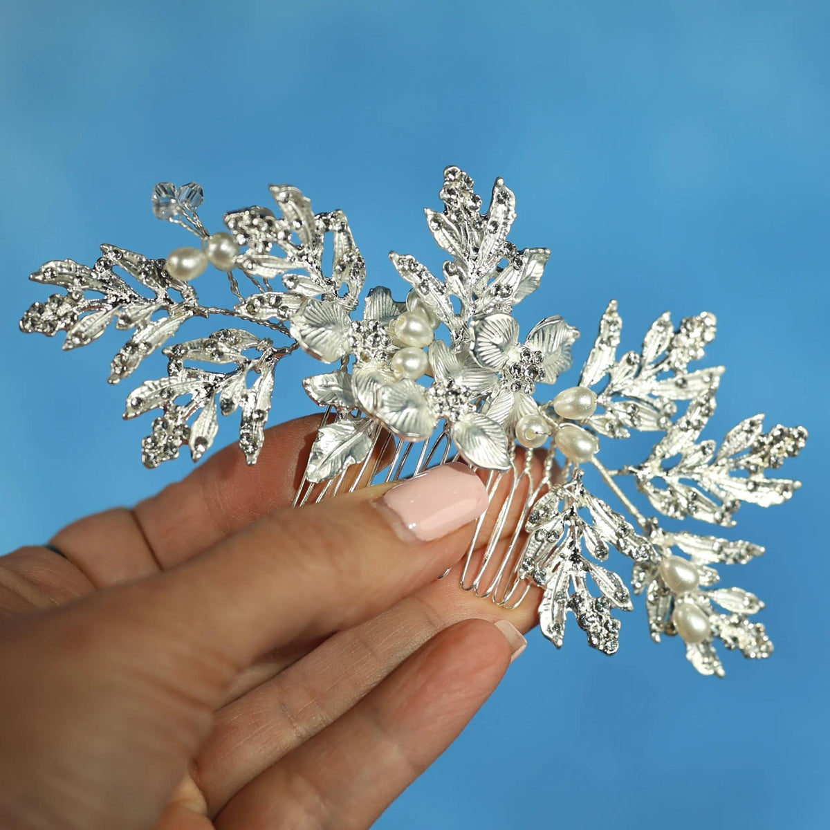 A hand holding a decorative hair accessory with white flowers and leaves against a blue background.