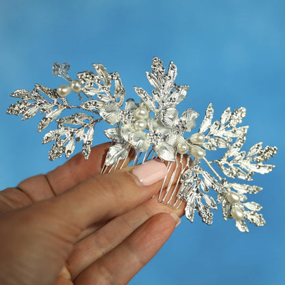 A hand holding a decorative hair accessory with white flowers and leaves against a blue background.