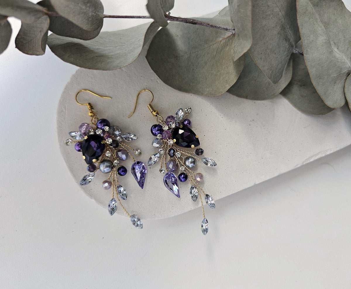 A pair of elegant earrings with purple and blue gemstones, hanging from gold hooks, placed on a white surface next to some green leaves.