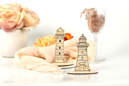 two wooden lighthouse models placed on a white surface, with a vase of pink roses and a glass of wine in the background.