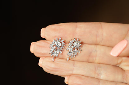 A close-up image of a person's hand holding a pair of diamond earrings against a dark background.
