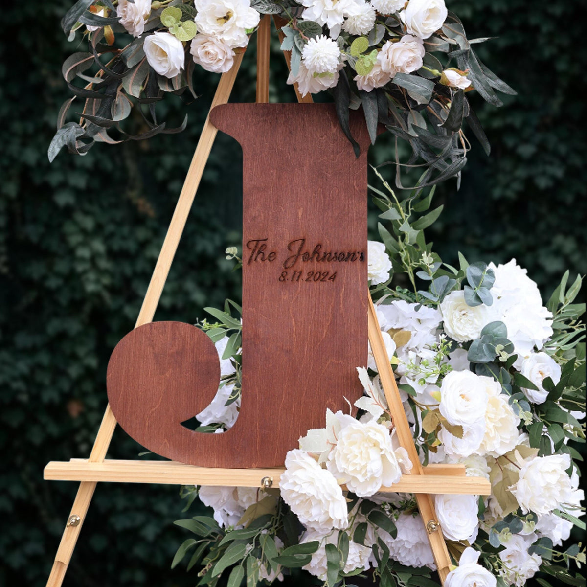 A wooden sign with the name "The Johnson's" and the date "8/11/2024" engraved on it, surrounded by a floral arrangement of white and green flowers.