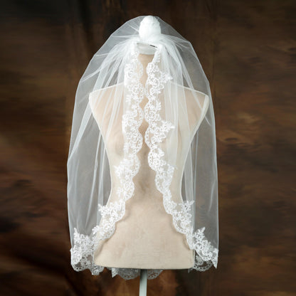 A white wedding veil with a delicate lace trim is displayed on a mannequin against a brown background.