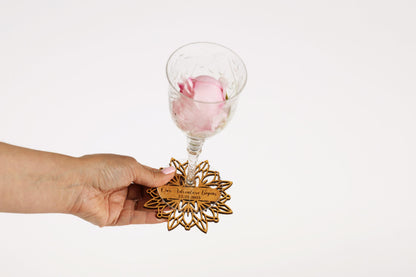 A hand holding a glass with a pink rose inside, and a wooden coaster with a nameplate attached to it.