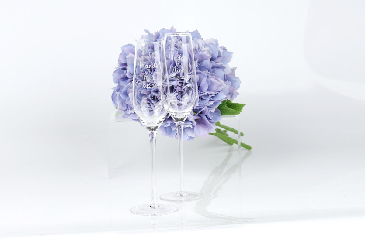 Two clear champagne glasses stand in front of a bouquet of purple hydrangeas, creating a visually appealing and elegant scene.