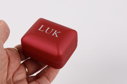 A hand holding a red jewelry box with the word "LUK" printed on it.
