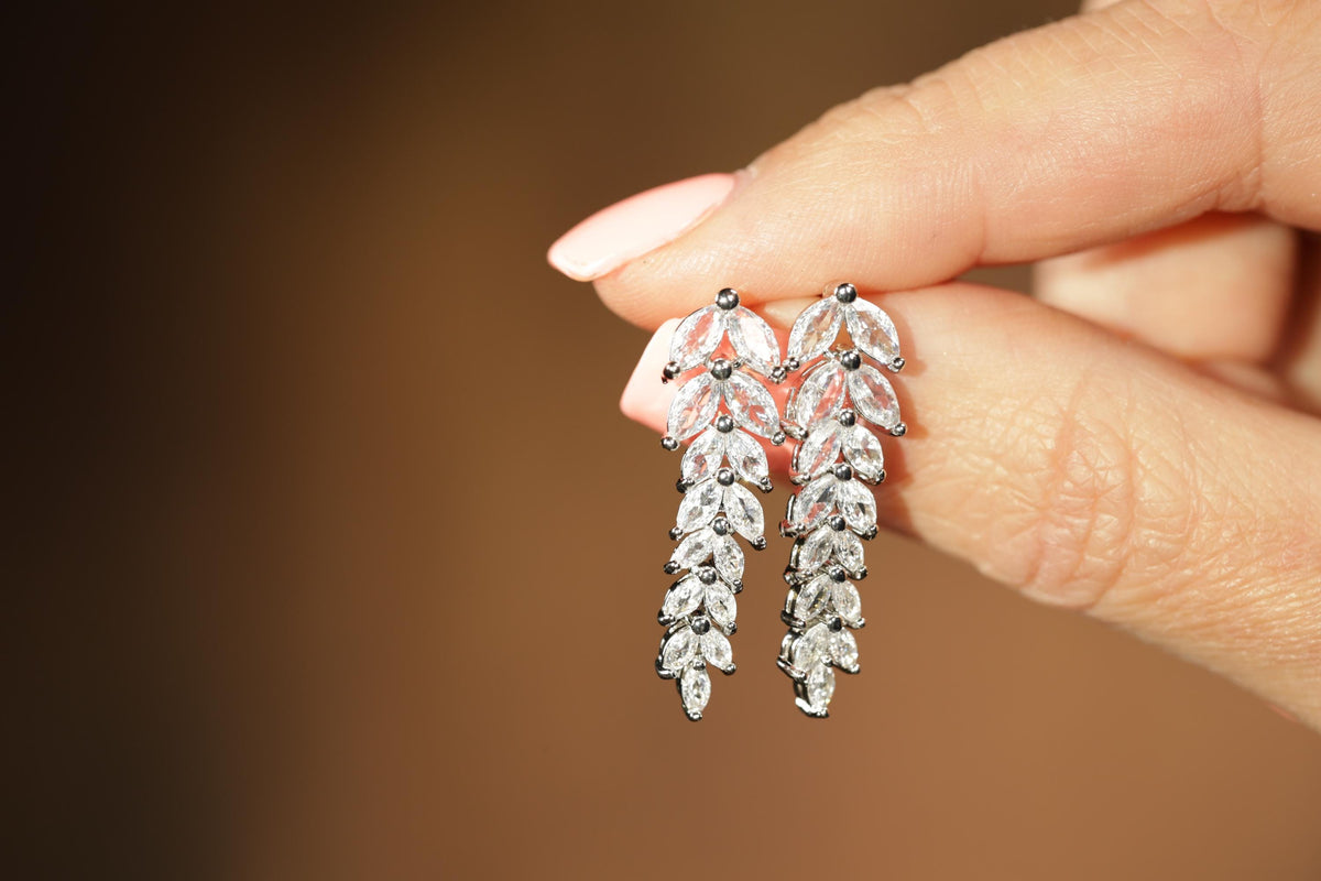 A hand holding a pair of elegant diamond earrings with intricate leaf-shaped designs.