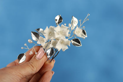 A hand holding a decorative hair clip with white flowers and silver leaves against a blue background.