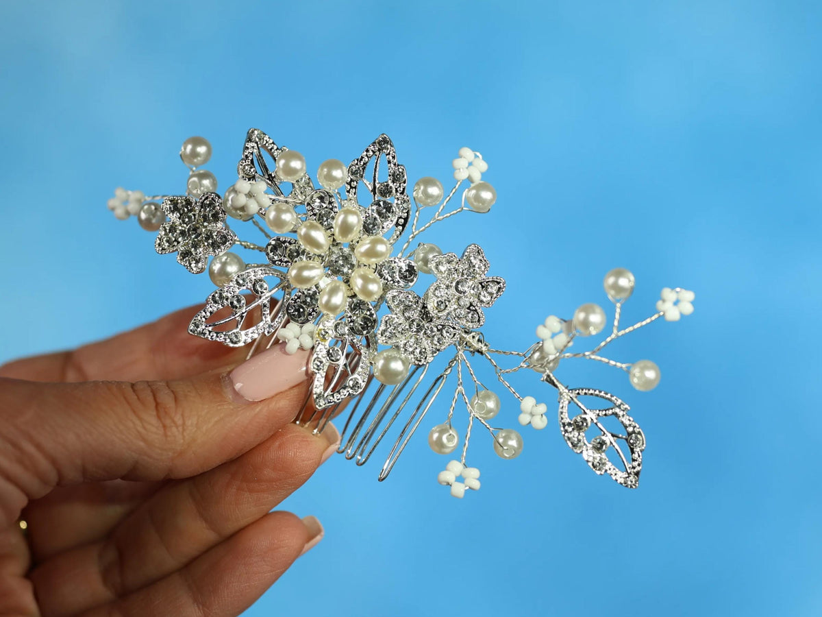 A hand holding a decorative hair accessory with white flowers and pearls against a blue background.