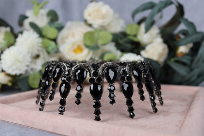 A black tiara with dangling black gemstones is displayed on a pink velvet background, with white flowers in the background.