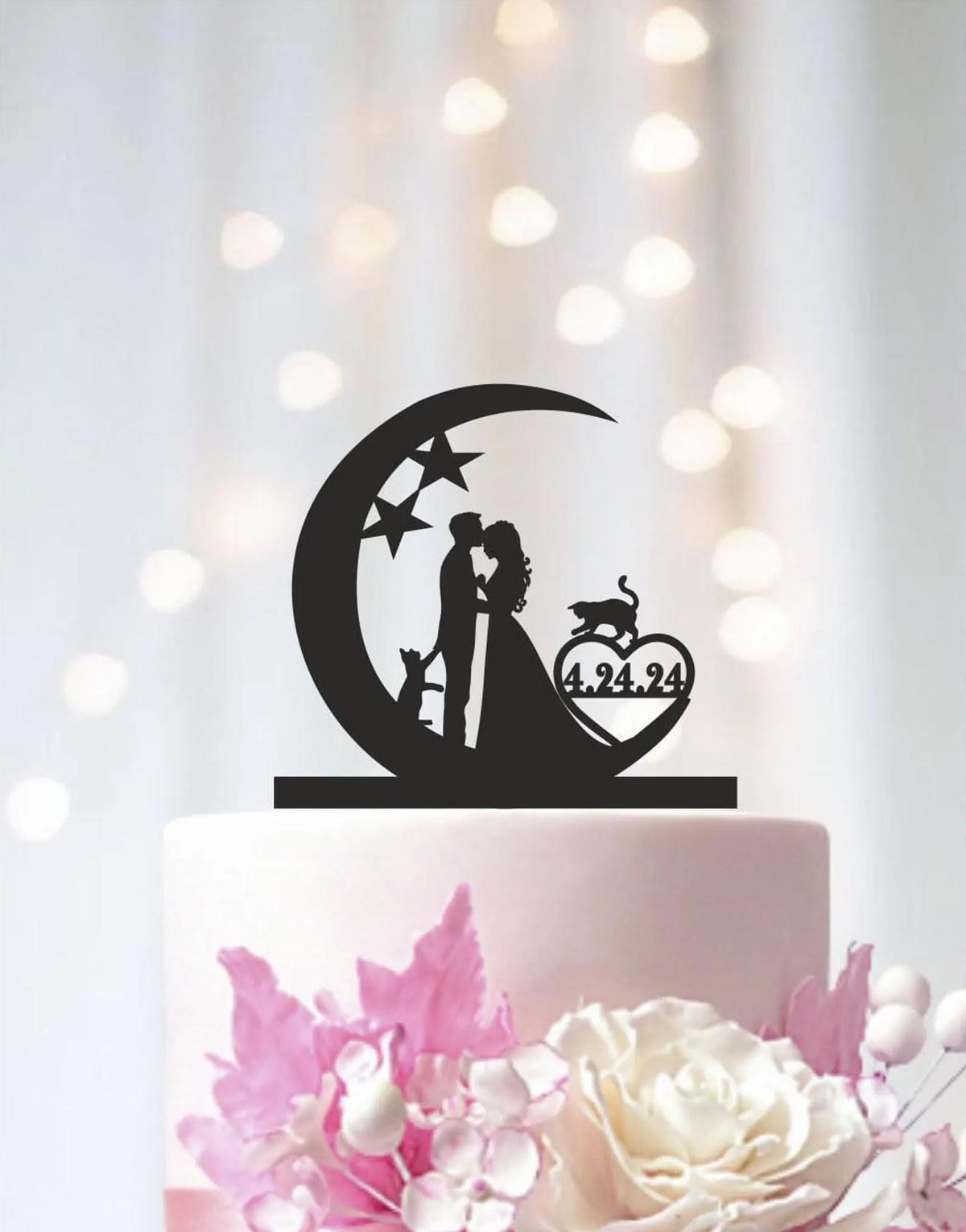 A black silhouette of a bride and groom kissing under a crescent moon on a wedding cake topper, with a heart-shaped decoration below.