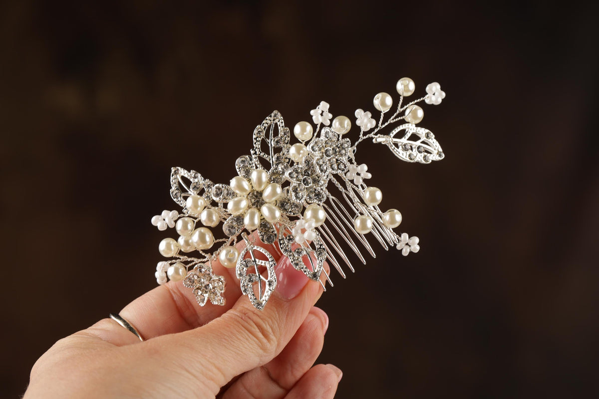 A hand holding a decorative hair accessory made of silver and white pearls and leaves.