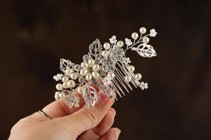 A hand holding a decorative hair accessory made of silver and white pearls and leaves.
