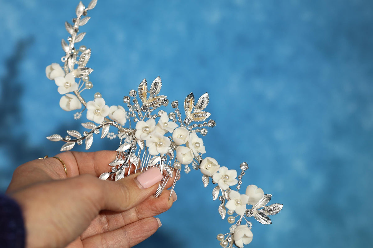 A hand holding a decorative hair accessory with white flowers and leaves against a blue background.