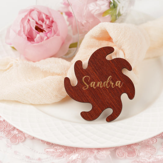 A wooden name tag with the name "Sandra" engraved on it, placed on a white plate with a pink flower and a white napkin.
